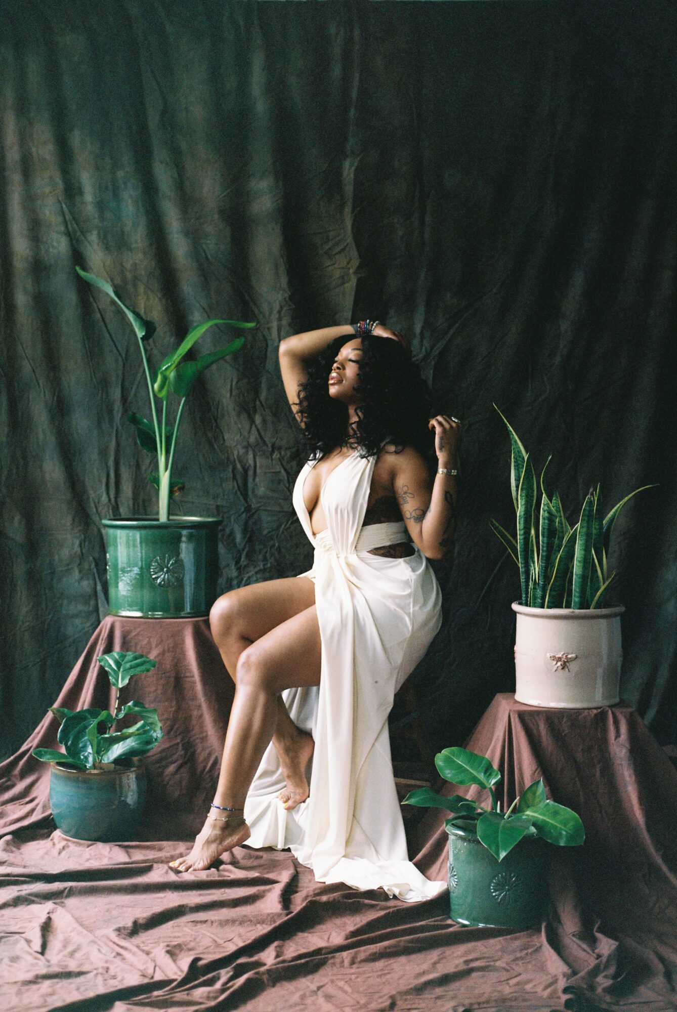 Woman sitting on a stool with plants around her, dark backdrop, wearing a white dress, one arm raised, head tilted back.