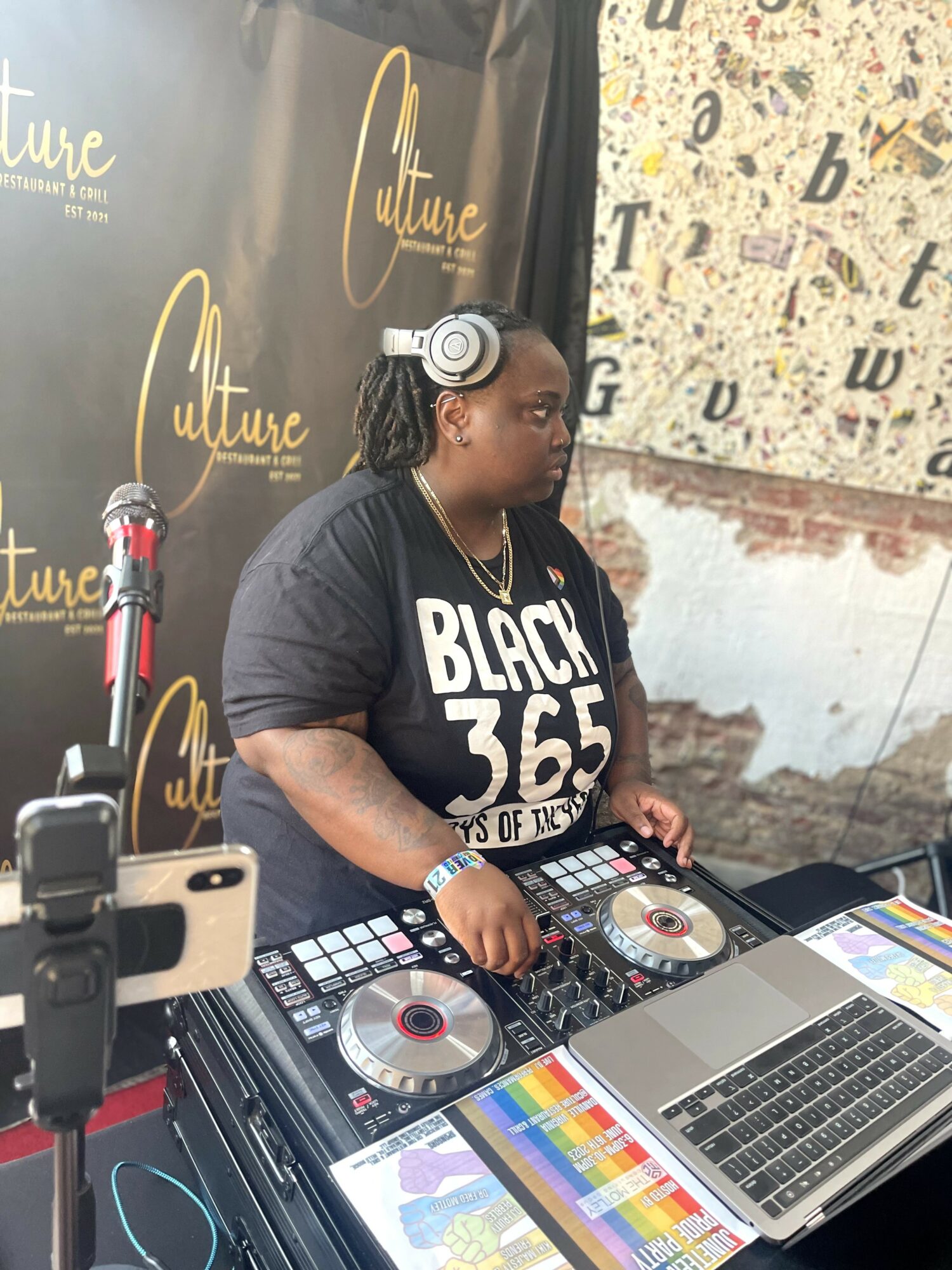 DJ woman with headphones operating a DJ controller and laptop at an event, with a black backdrop and patterned wall behind.