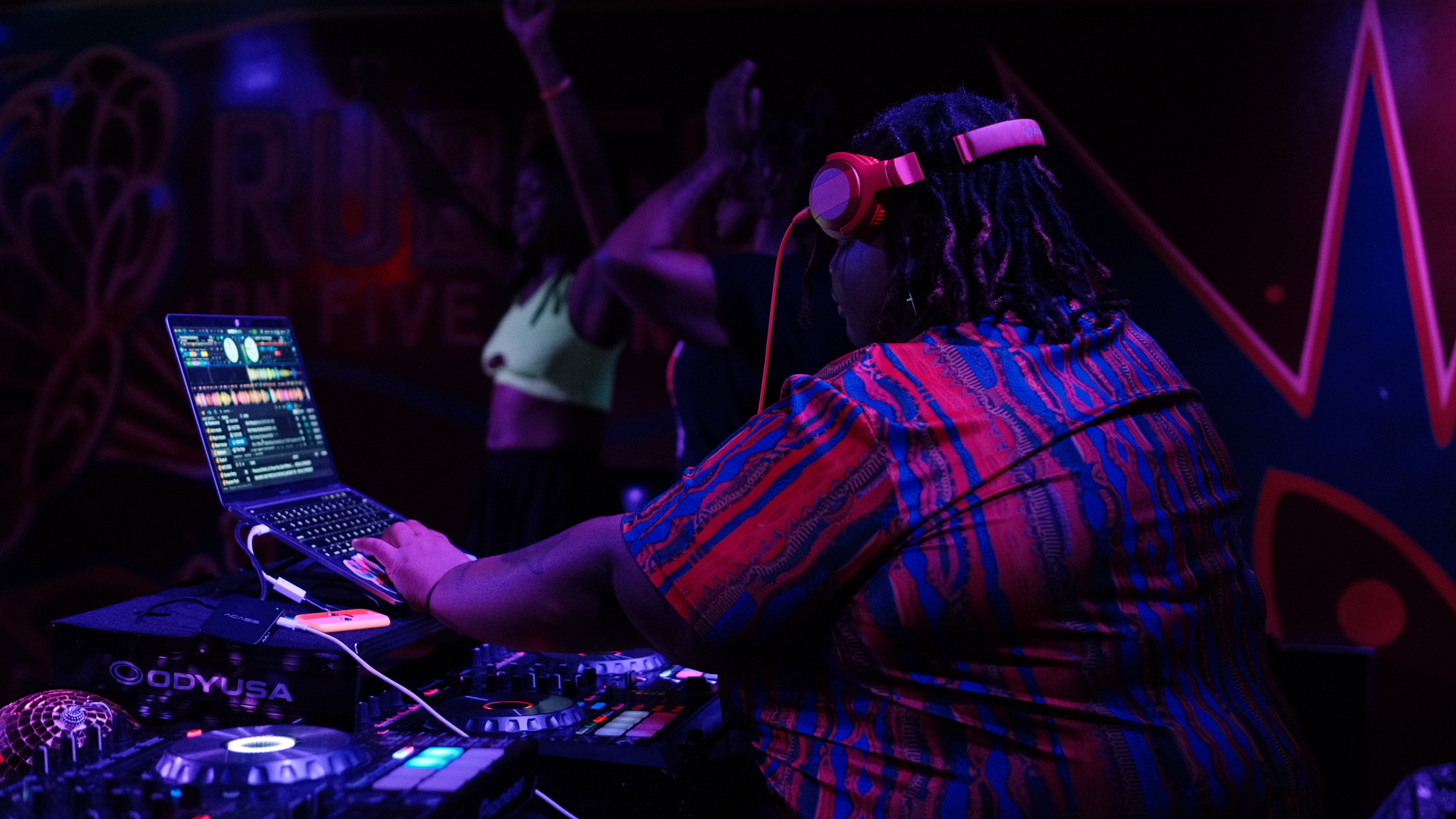 DJ wearing headphones operates mixing equipment in a dark, colorful club setting with neon lights.