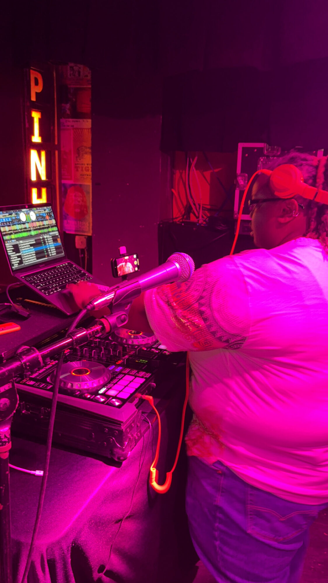 DJ wearing headphones operates equipment in a neon-lit nightclub with a sign reading 'PINK'.