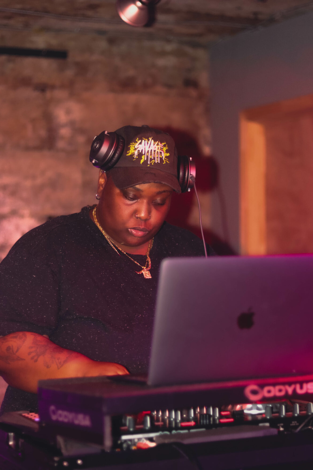 Woman DJ wearing headphones, focused on laptop, in a dimly lit room with brick wall background.