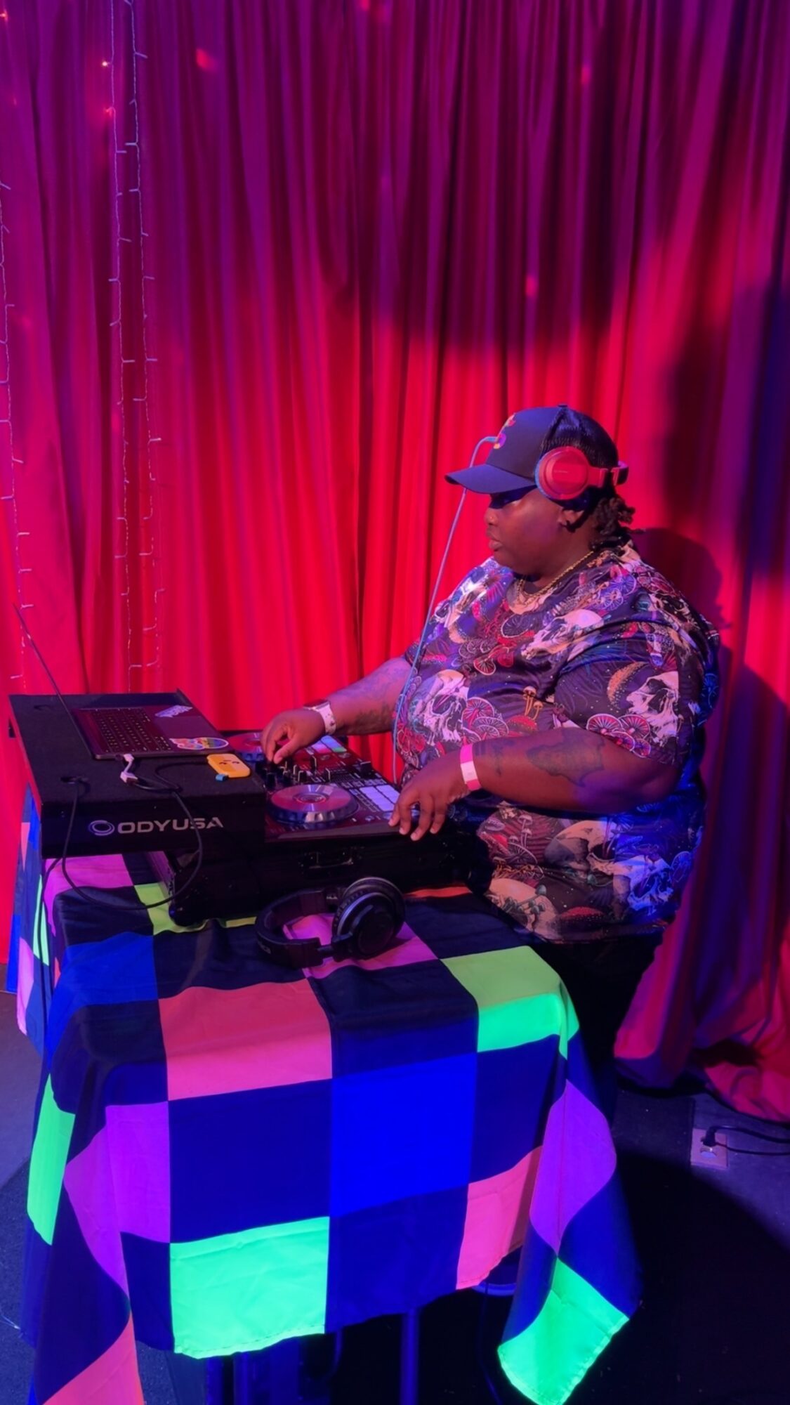 Person DJing with headphones, wearing a cap, in front of red curtains, using a laptop and DJ equipment, colorful tablecloth.