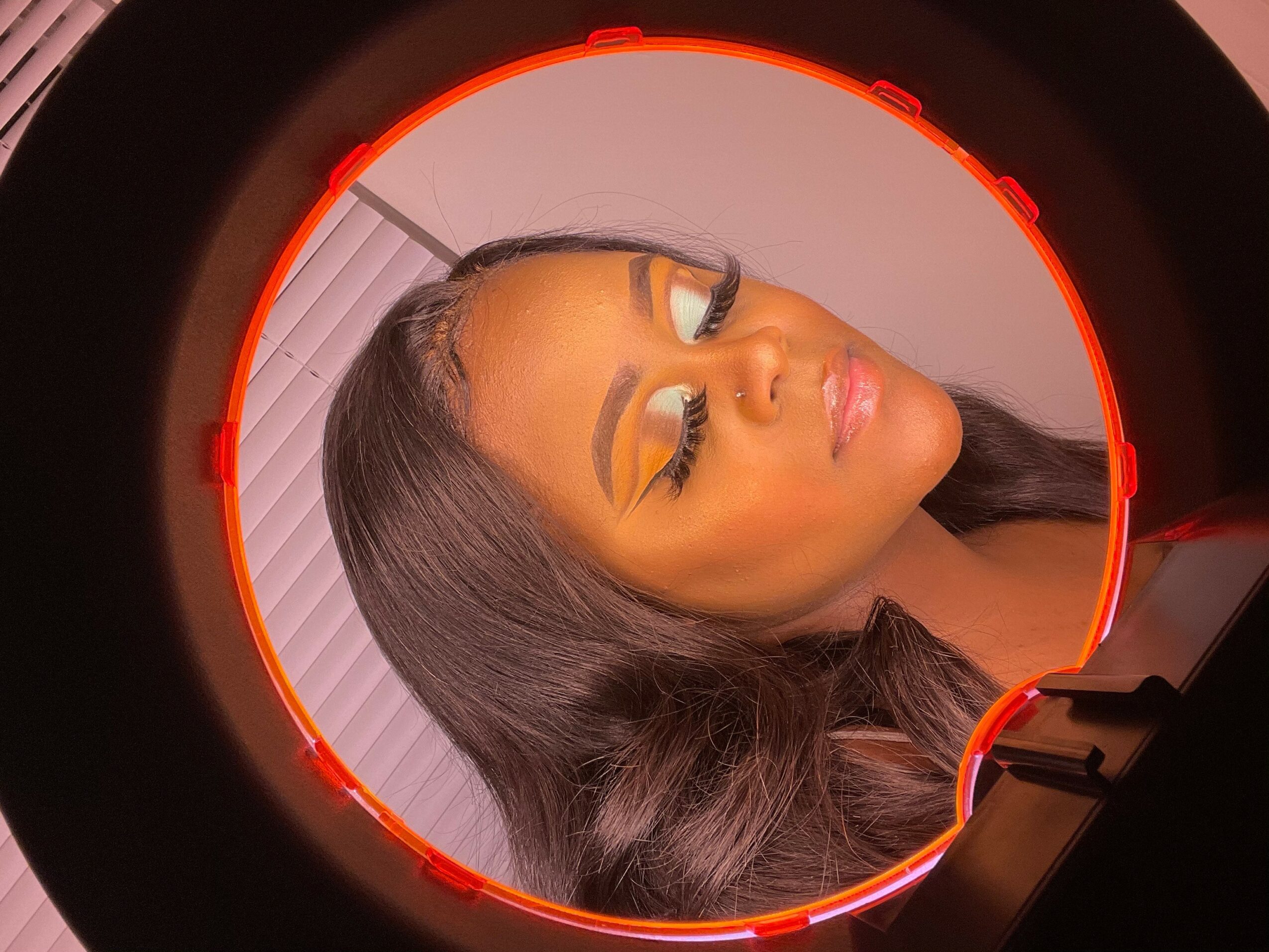 Woman with makeup and long hair, eyes closed, inside a circular device, background with vertical blinds.