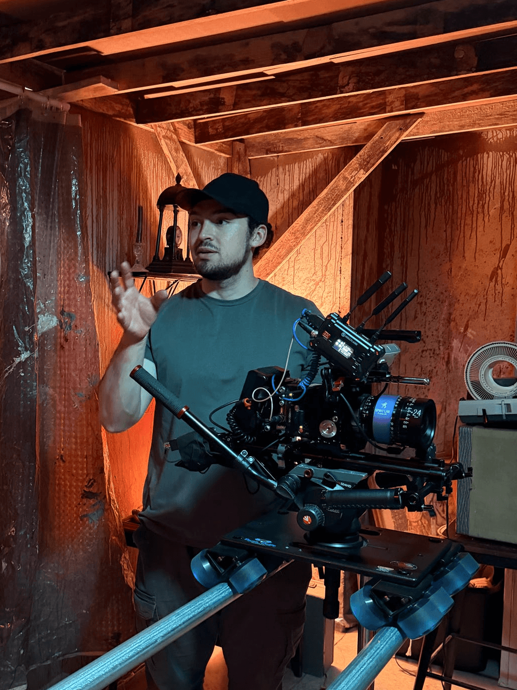 Man operating a camera rig in a wooden room, wearing a cap and casual clothing, with equipment and tools around him.