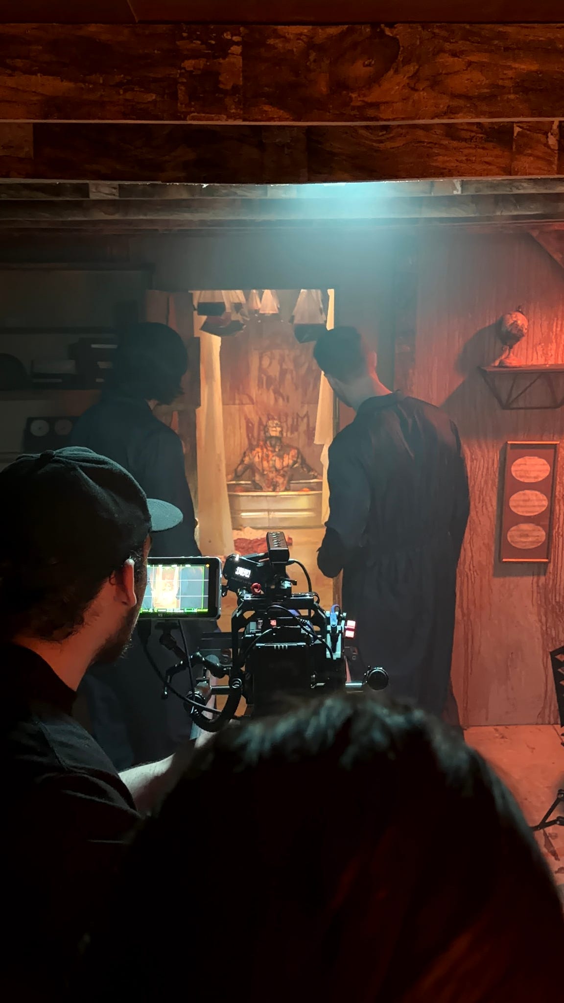People in a dimly lit room with a camera crew filming a scene, wooden walls, and a decorated background, 20 words.