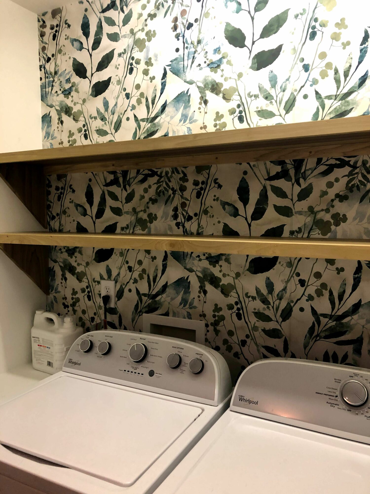 Laundry room with stacked washer and dryer units against a floral wallpapered wall, partially visible mirror on the left.