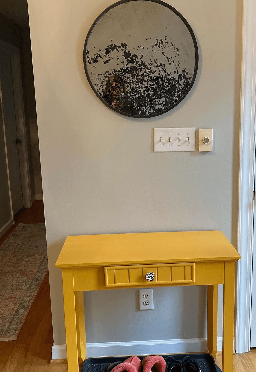 Round mirror above a yellow table with a drawer, wall with electrical outlets, and a doorway on the right.