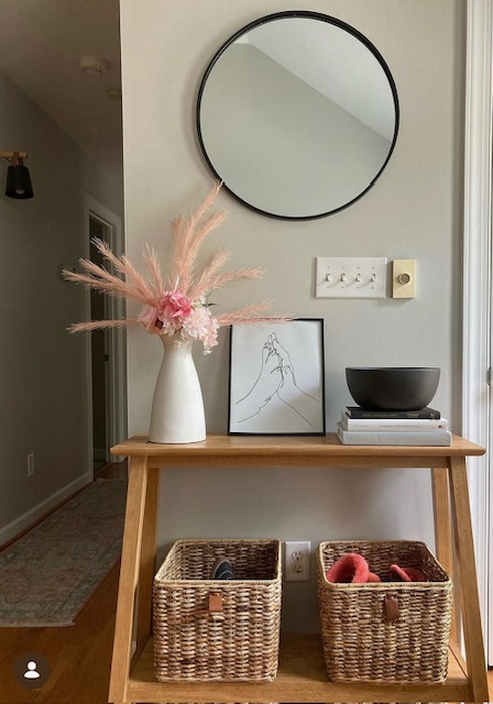 Decorative table with a vase of pink flowers, framed artwork, and a black bowl, against a wall with a round mirror.