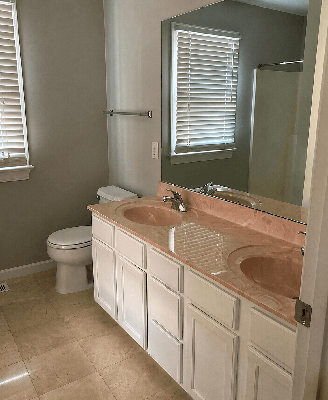 Bathroom with double sink vanity, mirror, toilet, window with blinds, and shower area, beige tiled floor, light green walls.