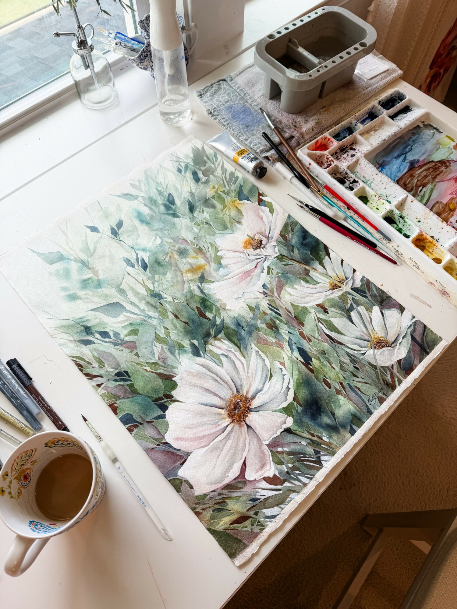 Watercolor painting of white flowers and green foliage on a table with art supplies nearby.