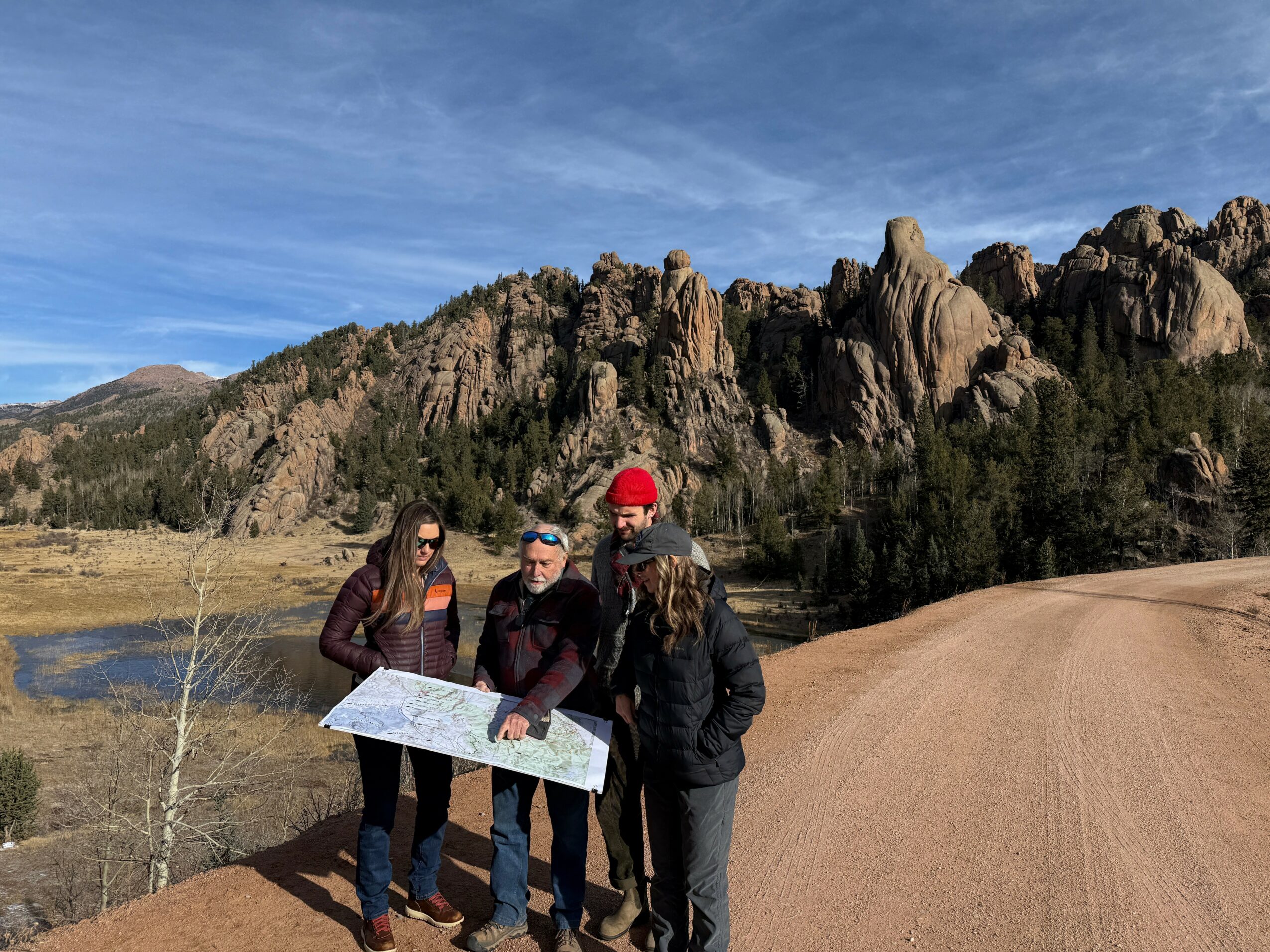 Four people stand on a dirt path near rocky hills and a body of water, looking at a map, under a blue sky.