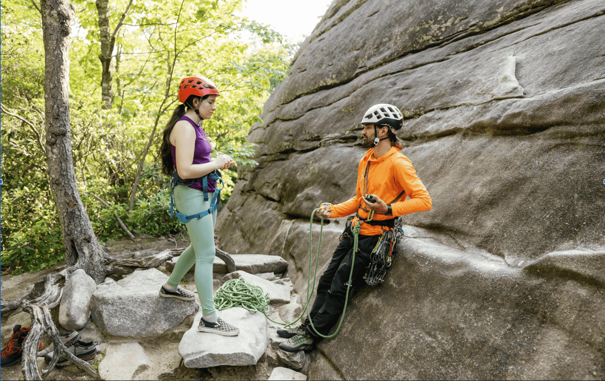 Two people with helmets and climbing gear near a large rock in a forested area.