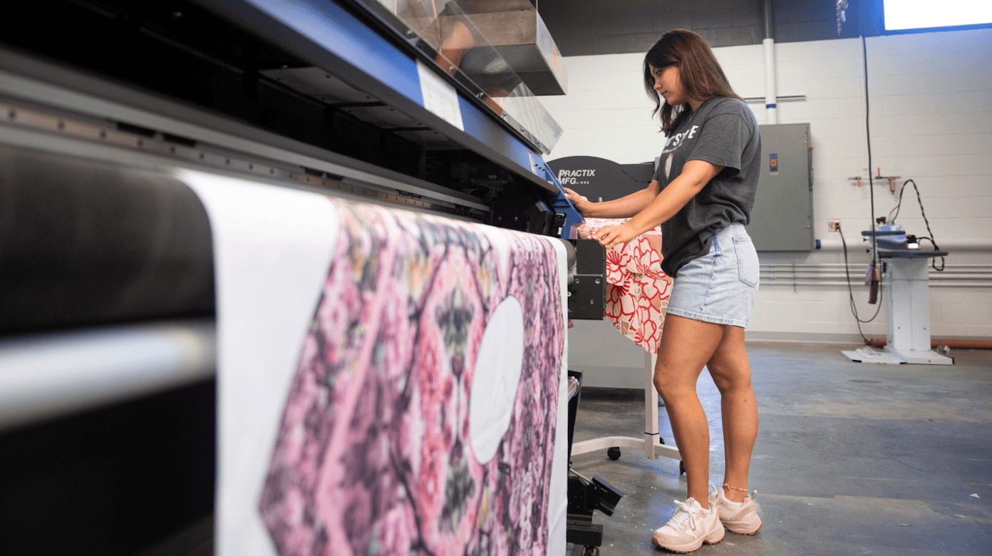 Young woman in shorts and t-shirt using a printing machine in a workshop, with fabric hanging from the machine.