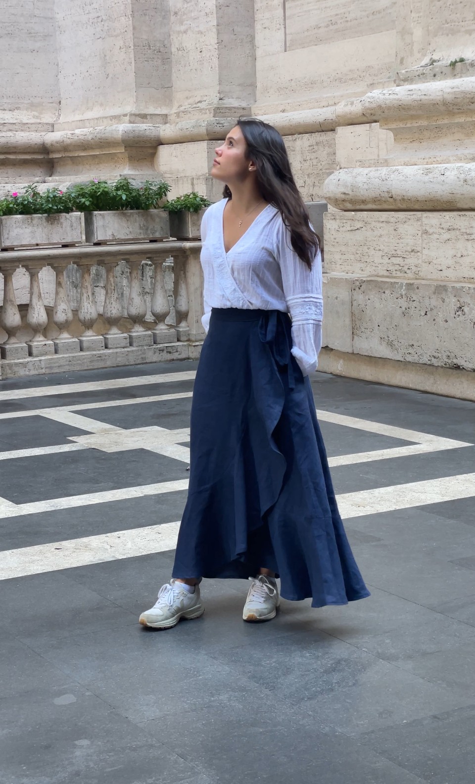 Woman standing outdoors near a stone building, wearing a white top and long dark skirt, looking to the side.