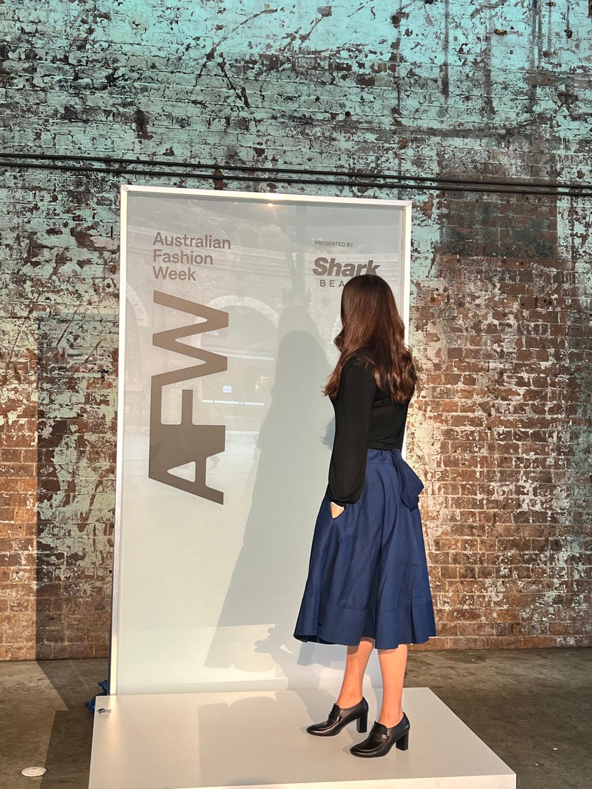 Woman with long hair in black top and blue skirt stands on a white platform facing a sign, brick wall background.
