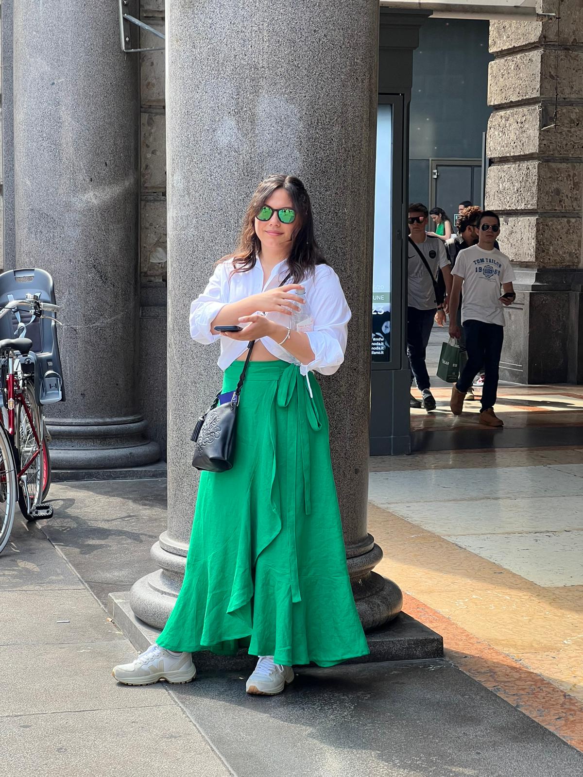 Woman in white shirt and green skirt standing next to a large column on a city sidewalk, with people in the background.