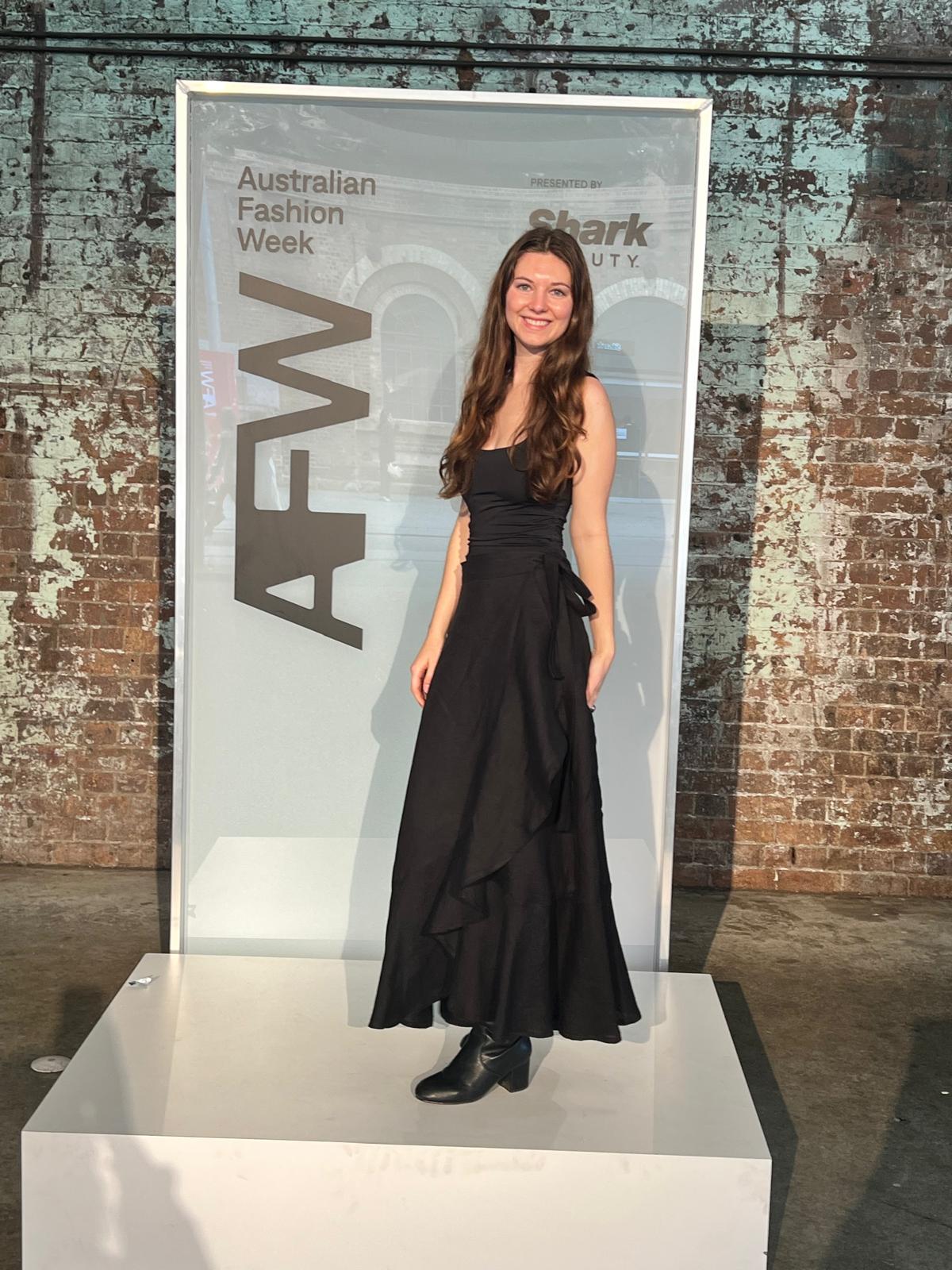 Young woman in a black dress and boots standing on a platform at Australian Fashion Week, brick wall background.