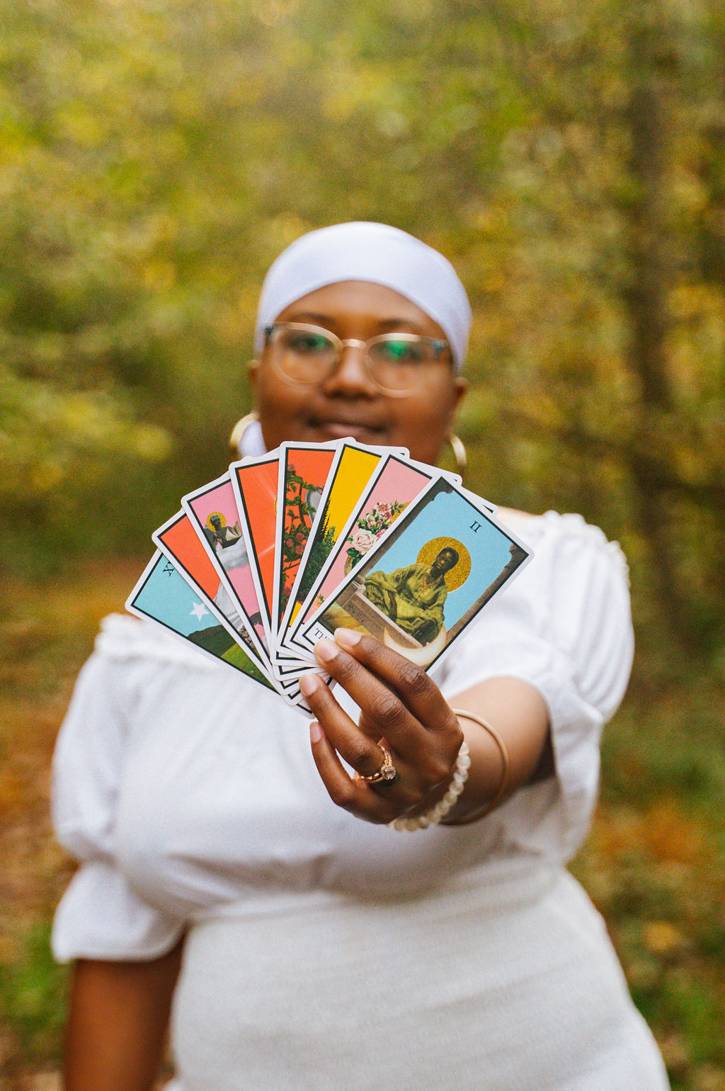 Person holding colorful cards outdoors with trees in the background, wearing glasses and a white headscarf.
