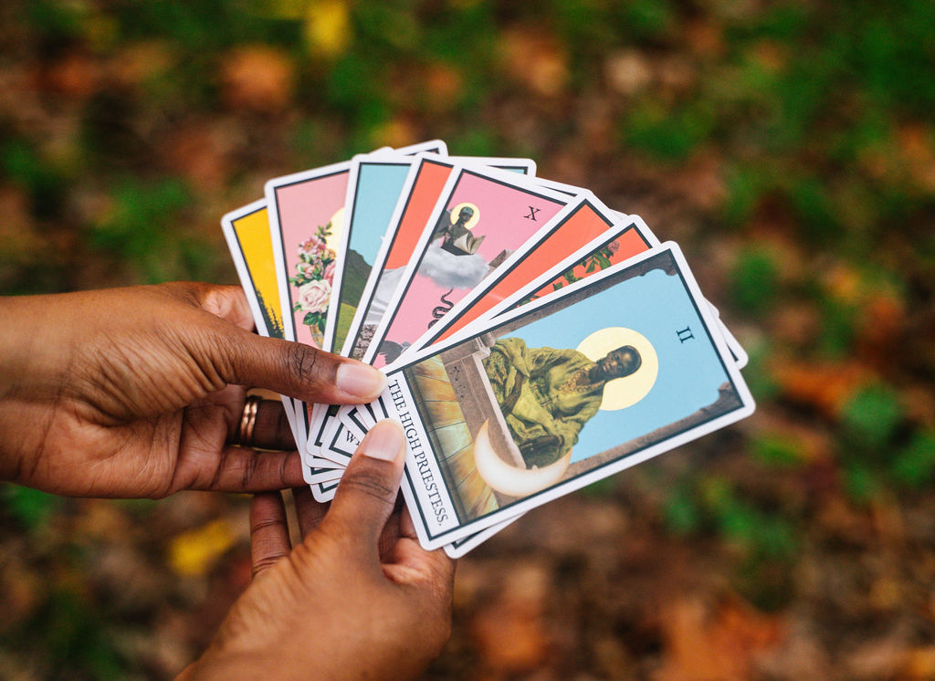 Person holding a fan of colorful tarot or playing cards outdoors with a blurred green and brown background.