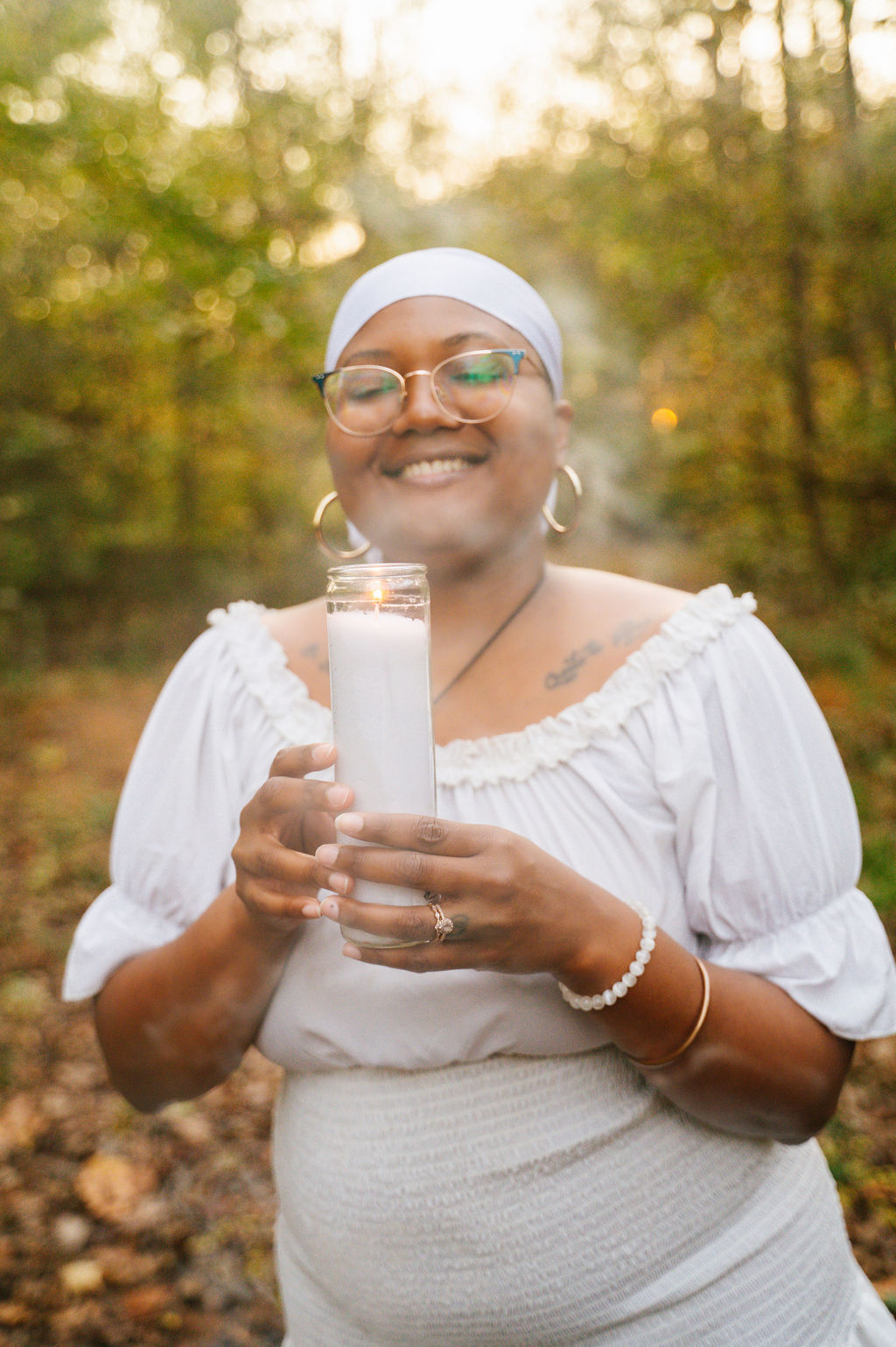 Smiling woman wearing glasses and a white headscarf holding a candle outdoors with trees in the background.
