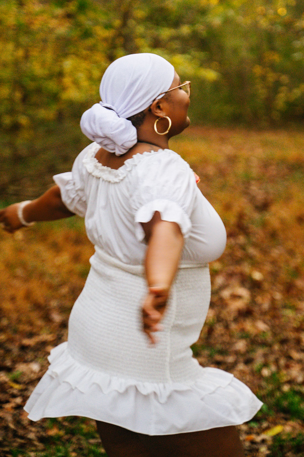 Woman in white dress with headscarf standing outdoors among autumn leaves, facing sideways, with trees in background.