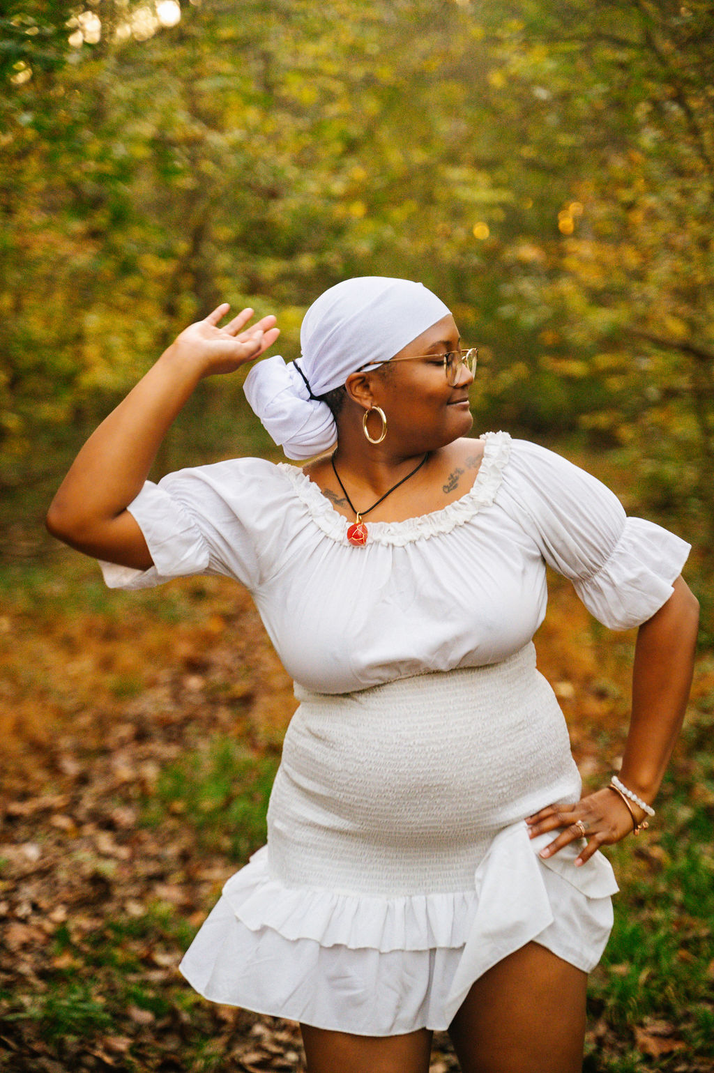 Woman in white dress with headscarf, glasses, and jewelry, standing outdoors in a forest during autumn, looking to the side.