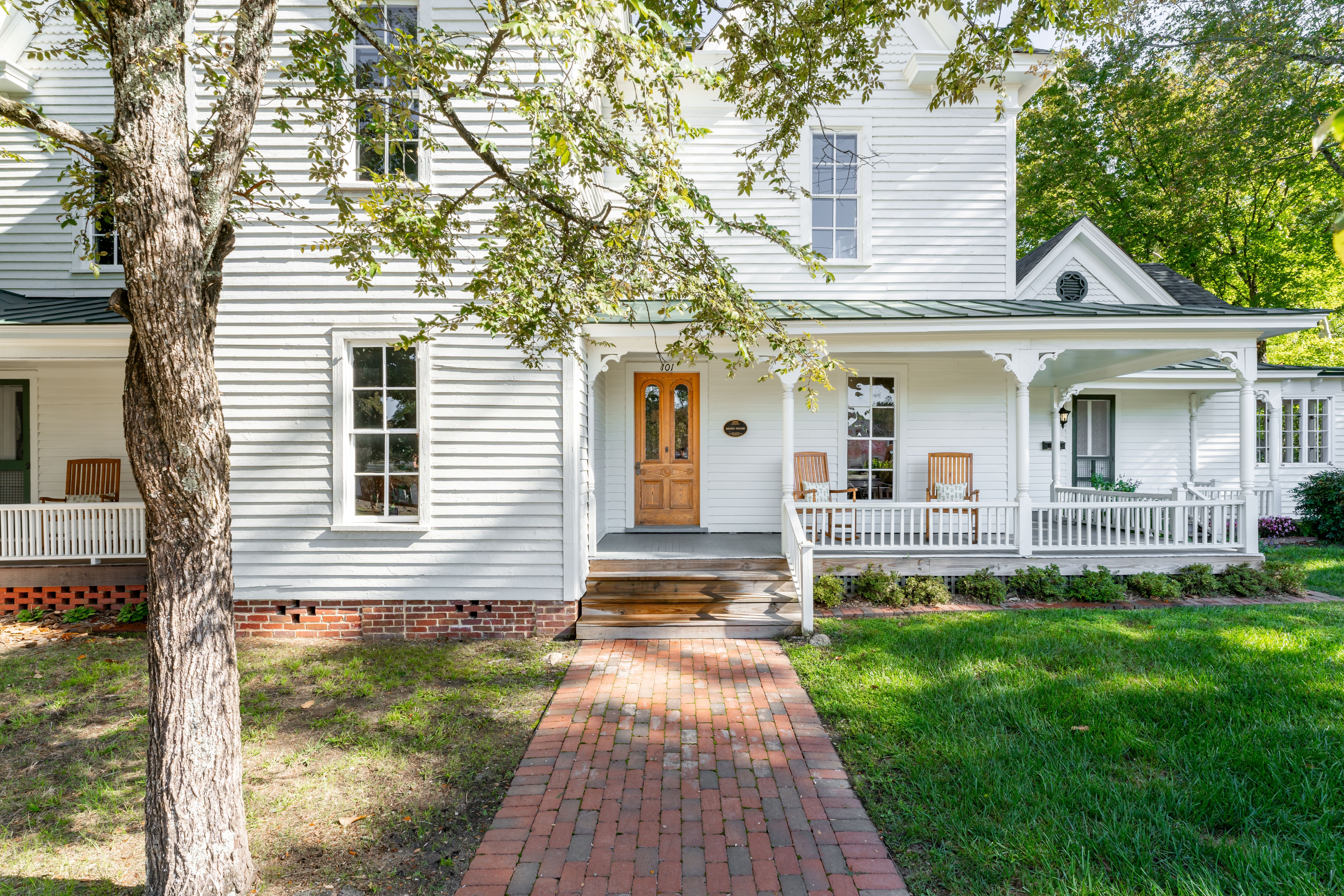White house with front porch, brick walkway, green lawn, and large tree in front yard.