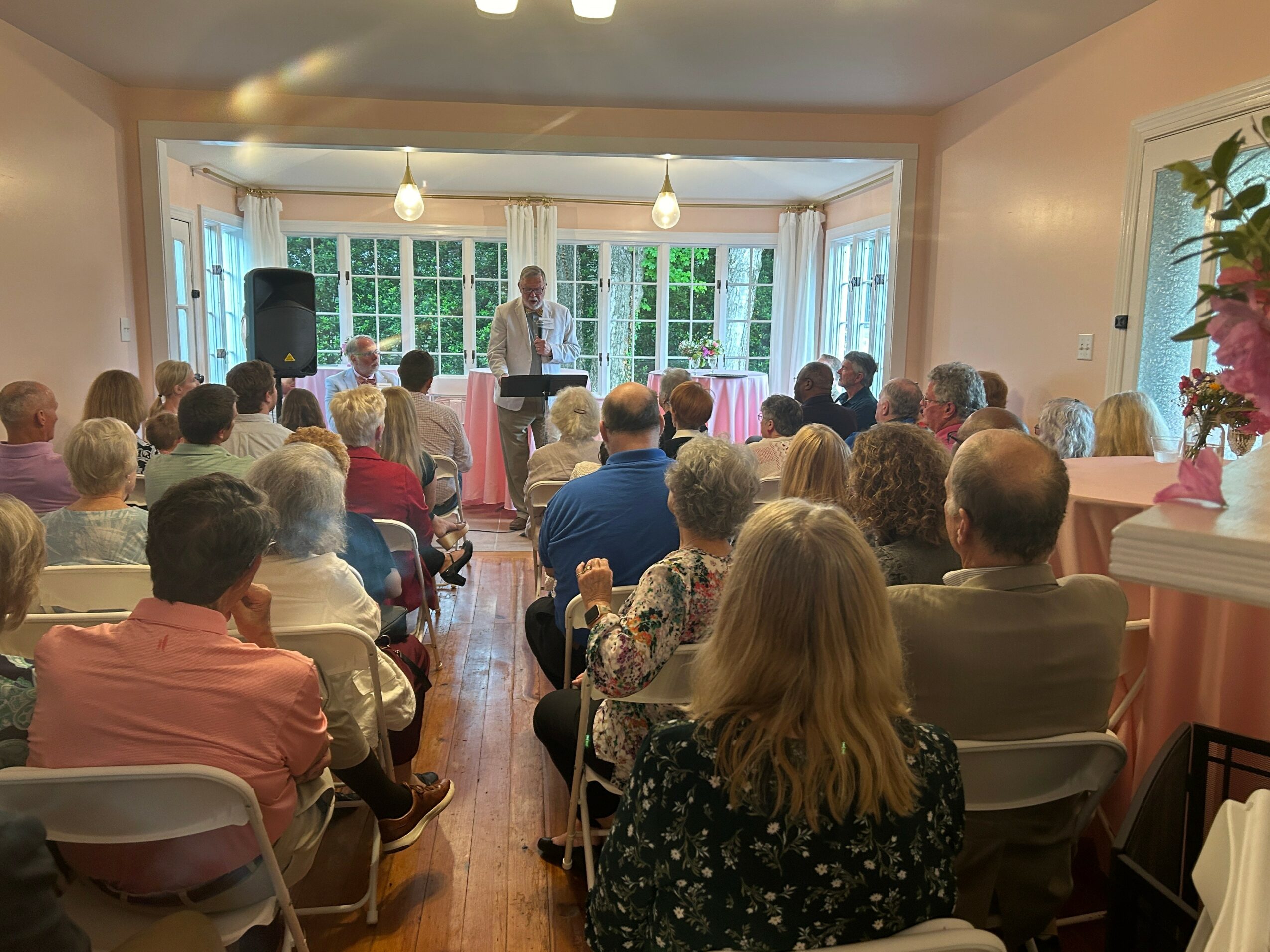 A speaker stands at a podium in front of a seated audience in a bright room with large windows and greenery outside.