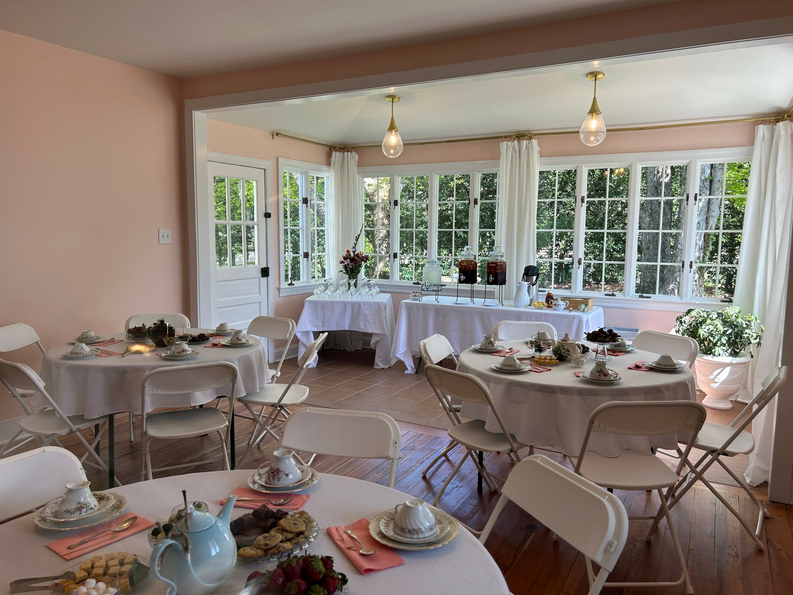 Dining room with round tables set with plates, cups, and napkins, large windows with white curtains, and a table with a floral centerpiece.