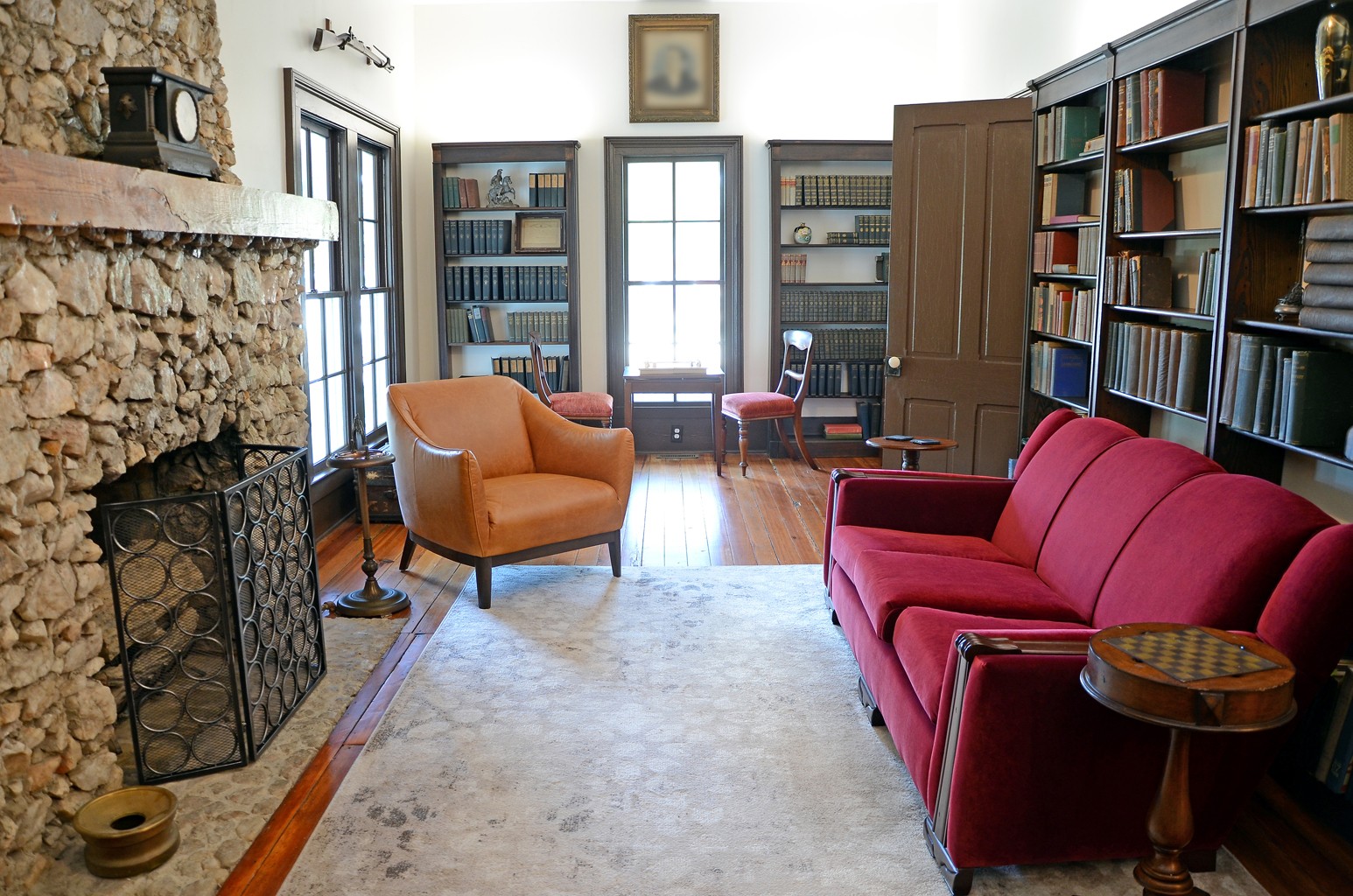 Living room with a stone fireplace, bookshelves, a red sofa, an orange armchair, and a window with sunlight.