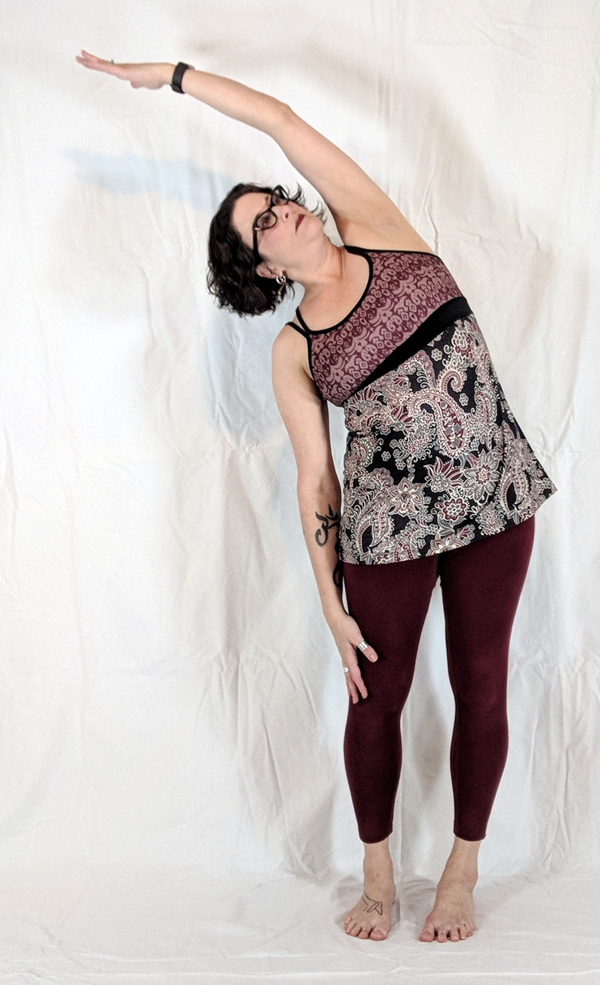 Person standing sideways with one arm raised above head, wearing glasses, patterned top, and maroon leggings, against a white background.