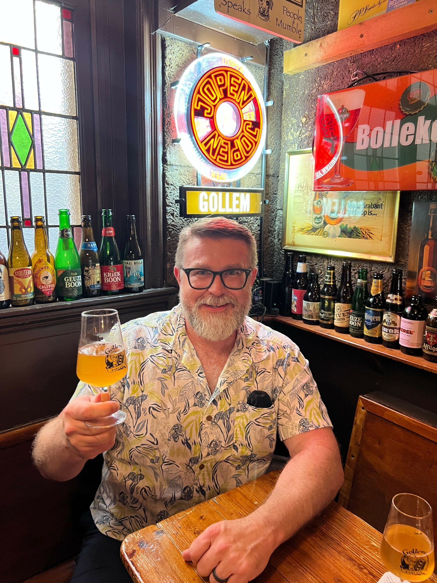Man with glasses in a patterned shirt holding a drink at a bar with bottles and signs behind him.