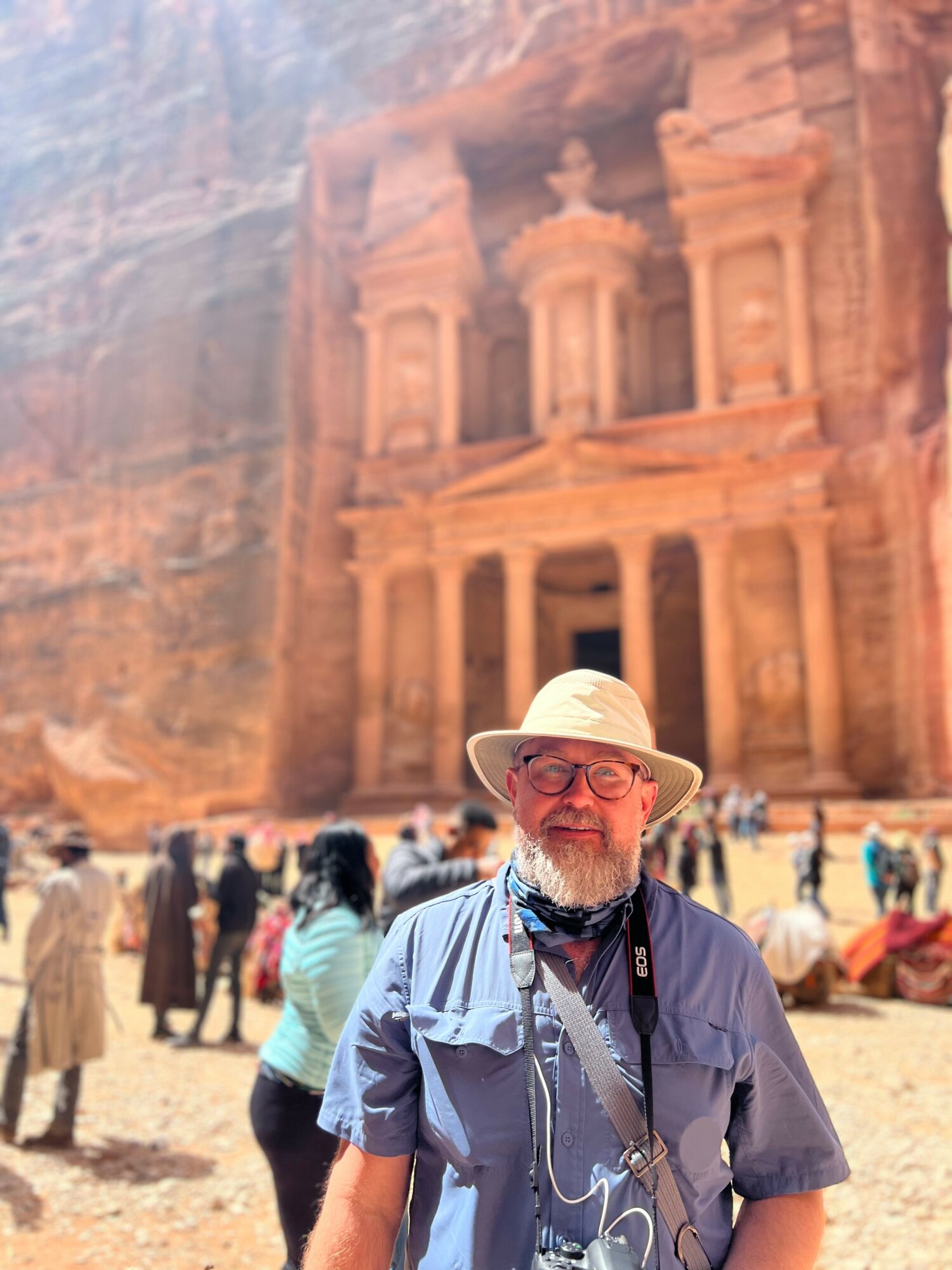 Older man with glasses, wearing a wide-brimmed hat and blue shirt, standing outdoors with a historic rock-cut structure in background.
