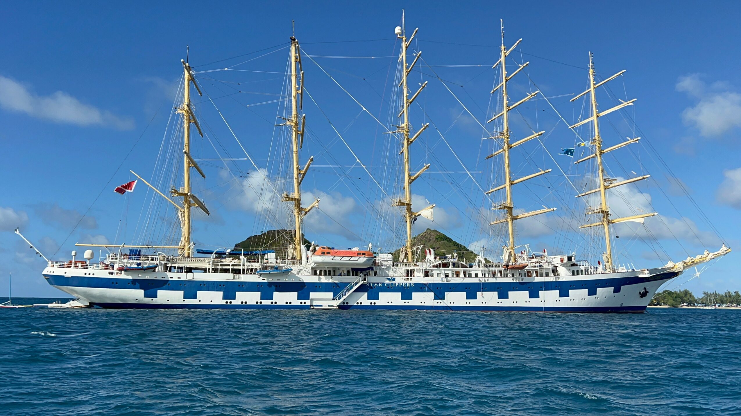 A large sailing ship with four tall masts and white and blue hull, sailing on the ocean with a small island in background.