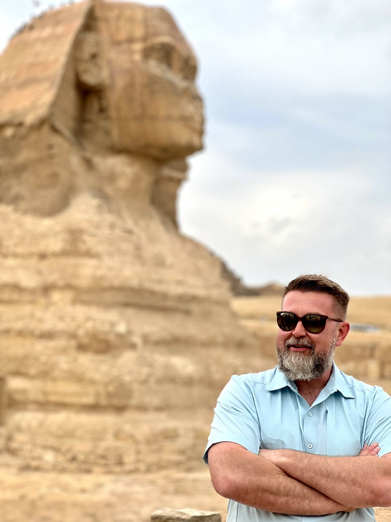 Man with sunglasses and a beard stands with arms crossed in front of the Sphinx in Egypt.