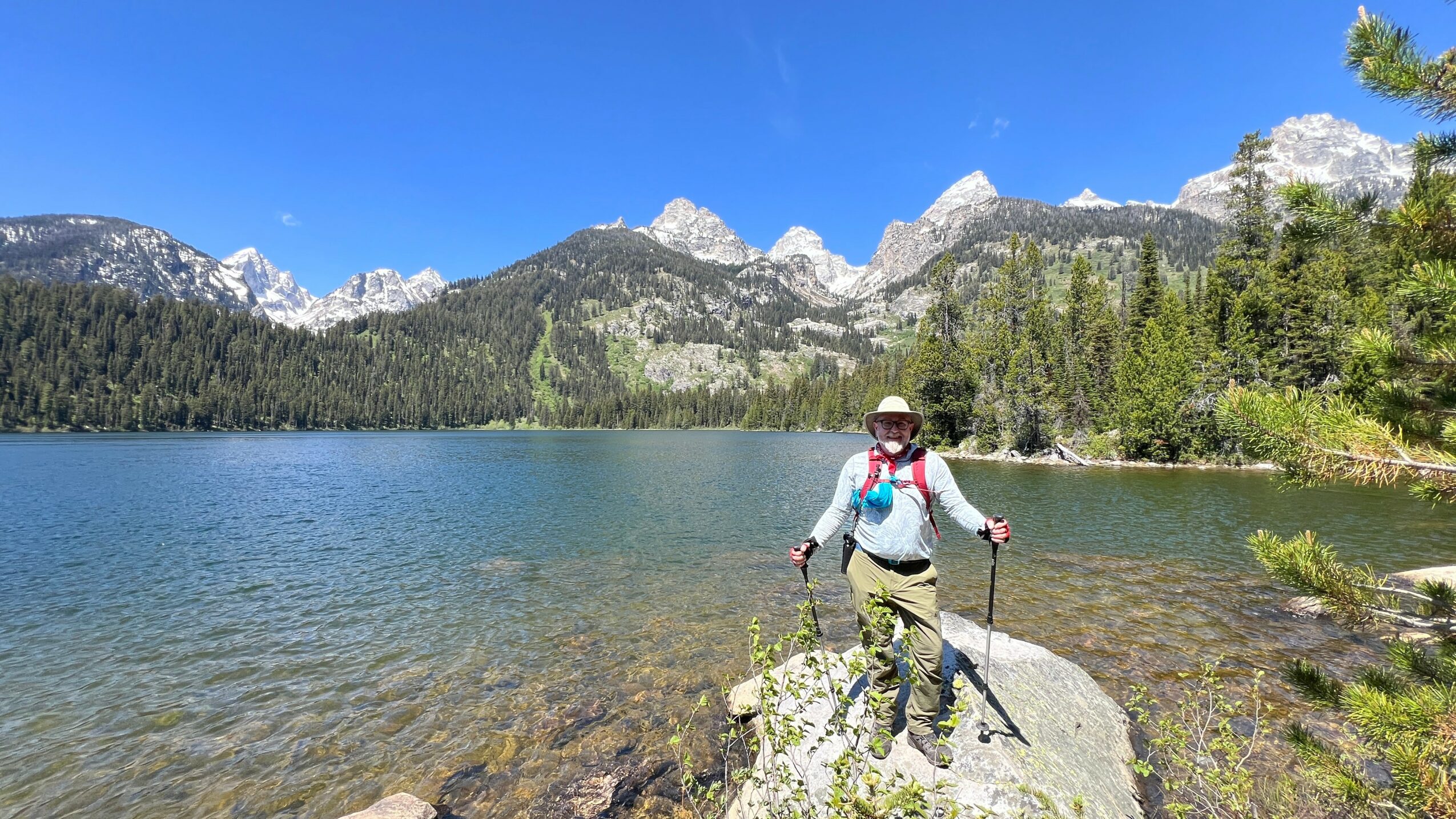 Person standing on a rock by a lake with mountains and trees in the background, holding hiking poles.