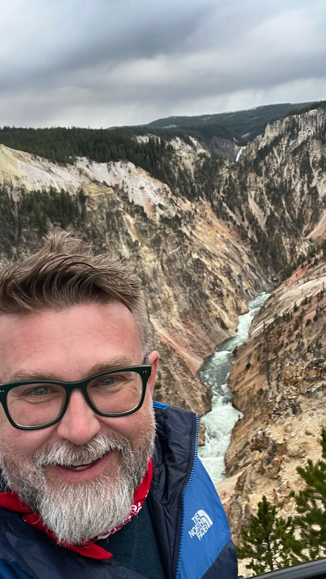 A man with glasses and a beard smiling in front of a canyon with a river and cliffs.