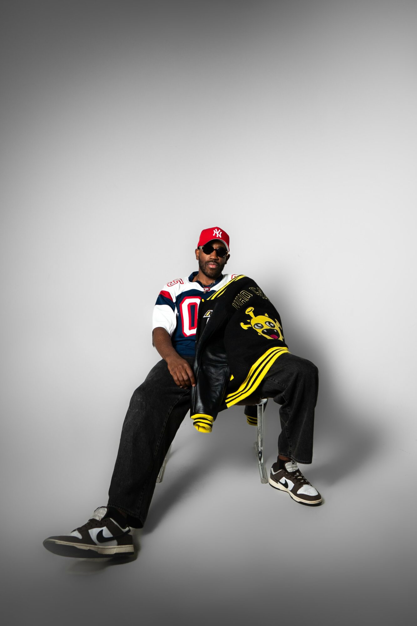 Person sitting on a chair against a plain background, wearing a sports jersey, cap, and sneakers.