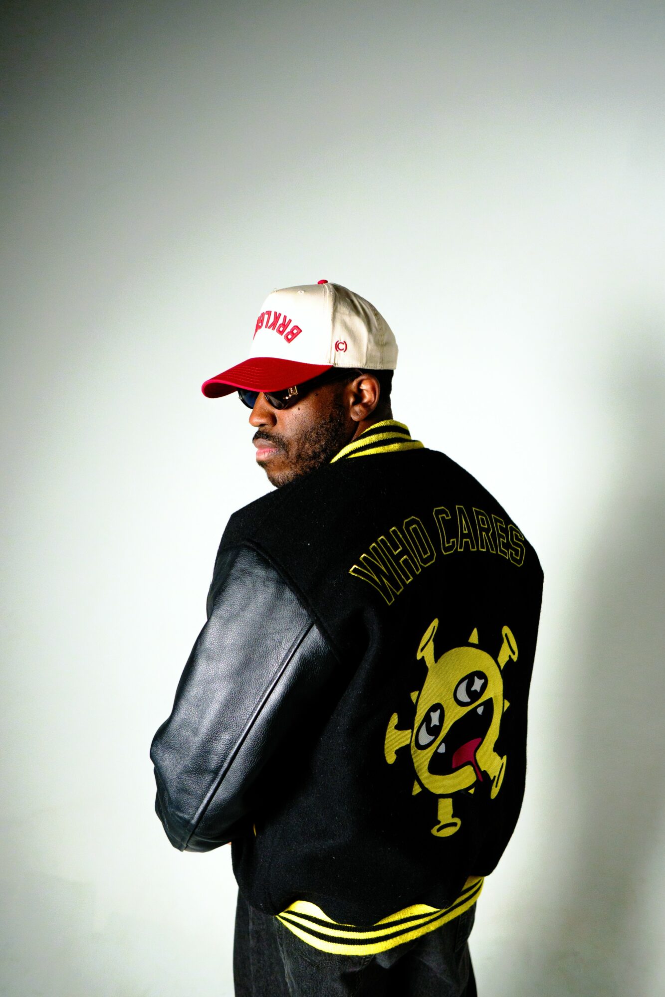 Man wearing a black jacket with a yellow cartoon character on the back, red and white cap, and sunglasses, facing sideways.