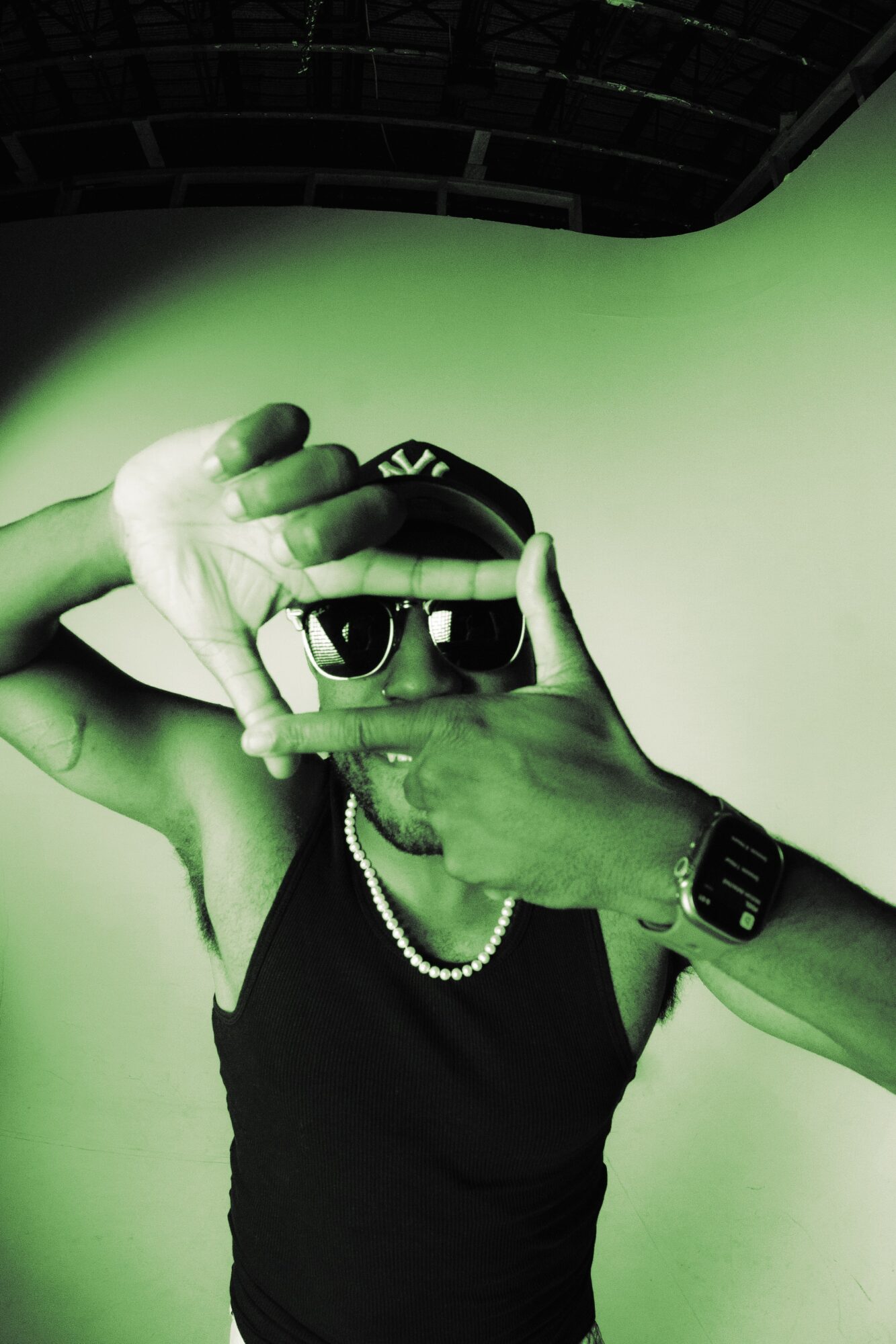 Person wearing sunglasses, a black tank top, and a pearl necklace making a frame with hands, against a green screen background.