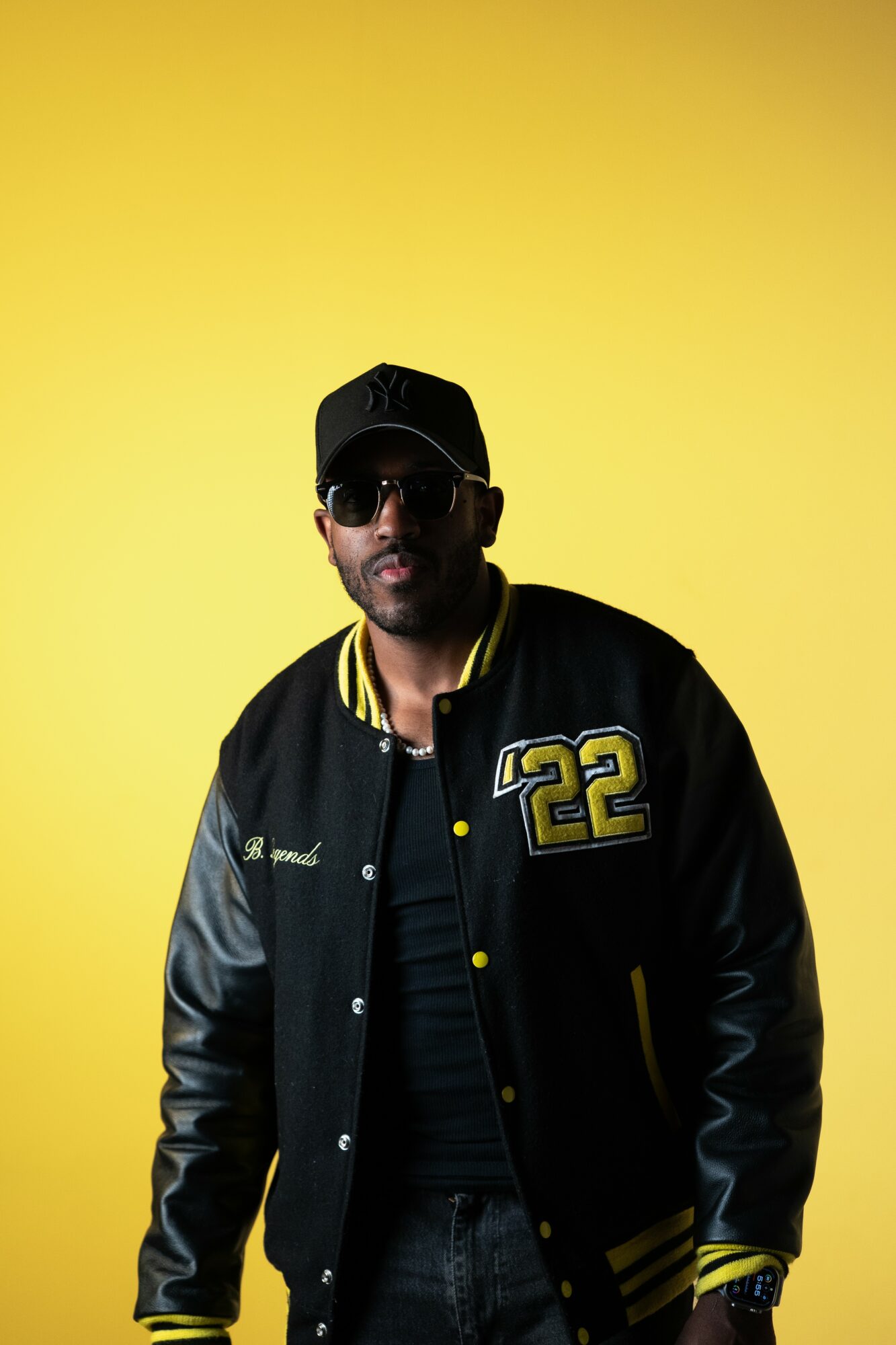 Man wearing sunglasses, black cap, and a black varsity jacket with yellow accents and the number 22, standing against yellow background.