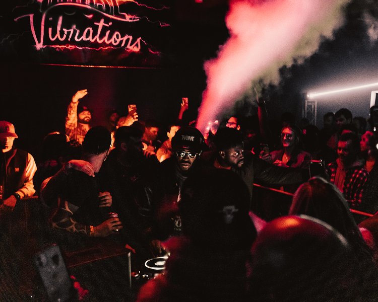 Crowd of people dancing and socializing in a dark club with pink neon sign reading 'Vinations' and smoke effects.