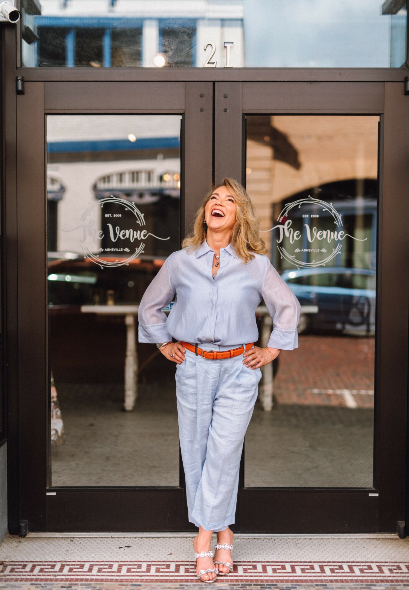 Woman standing in front of glass door, smiling, wearing light blue outfit, with reflections of buildings and street behind her.