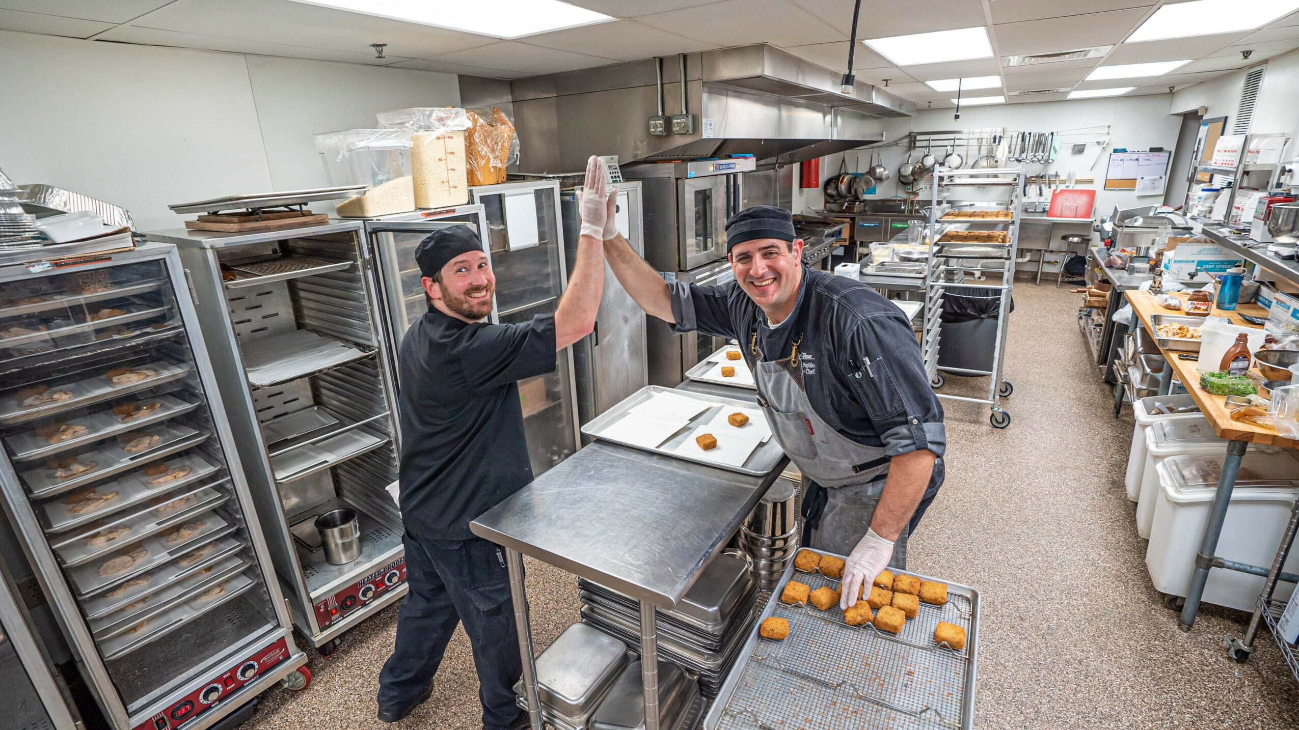 Two bakers in a commercial kitchen high-five over a tray of baked goods, surrounded by shelves and kitchen equipment.