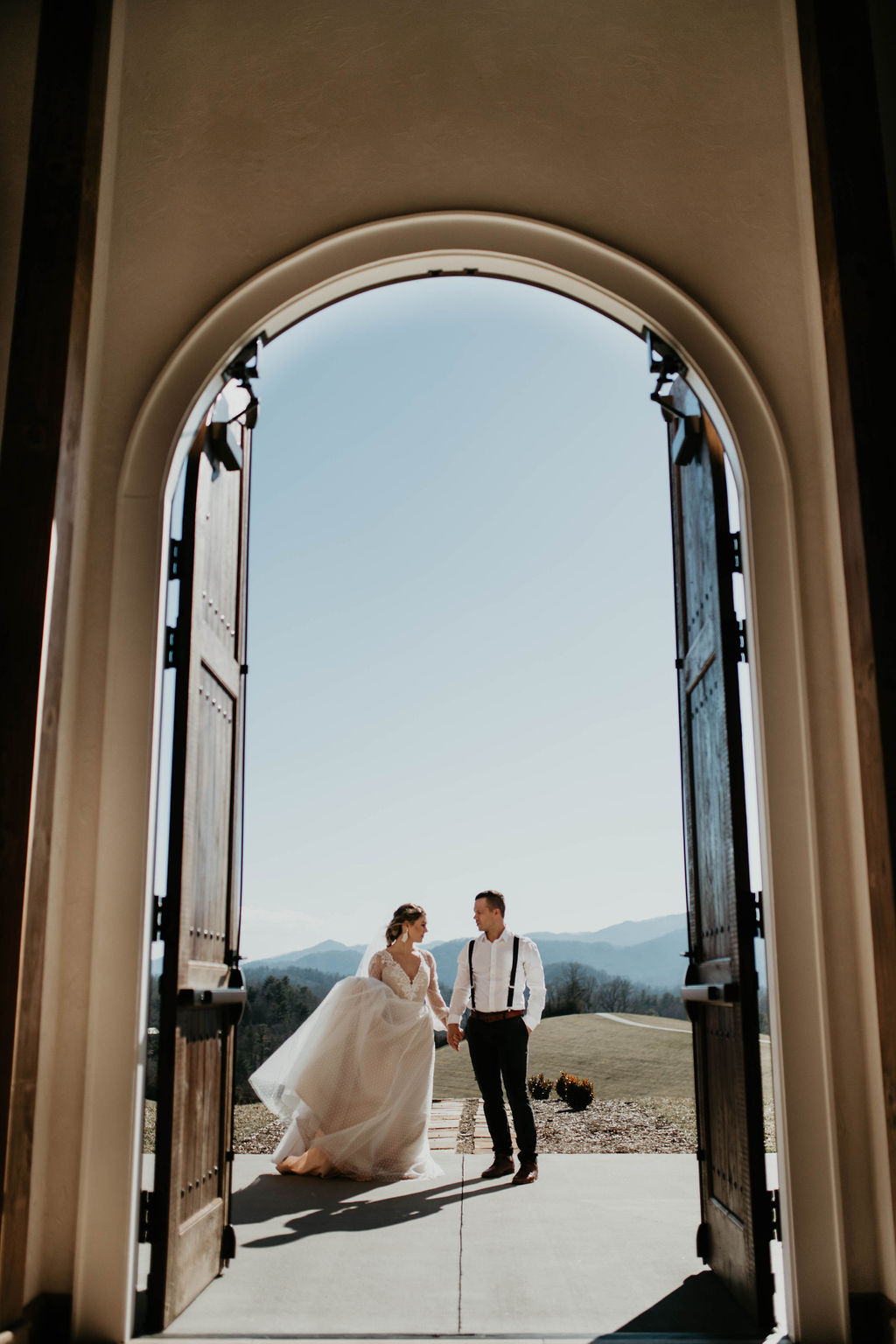 A bride and groom stand outside a building under an open arched doorway with a scenic landscape in the background.