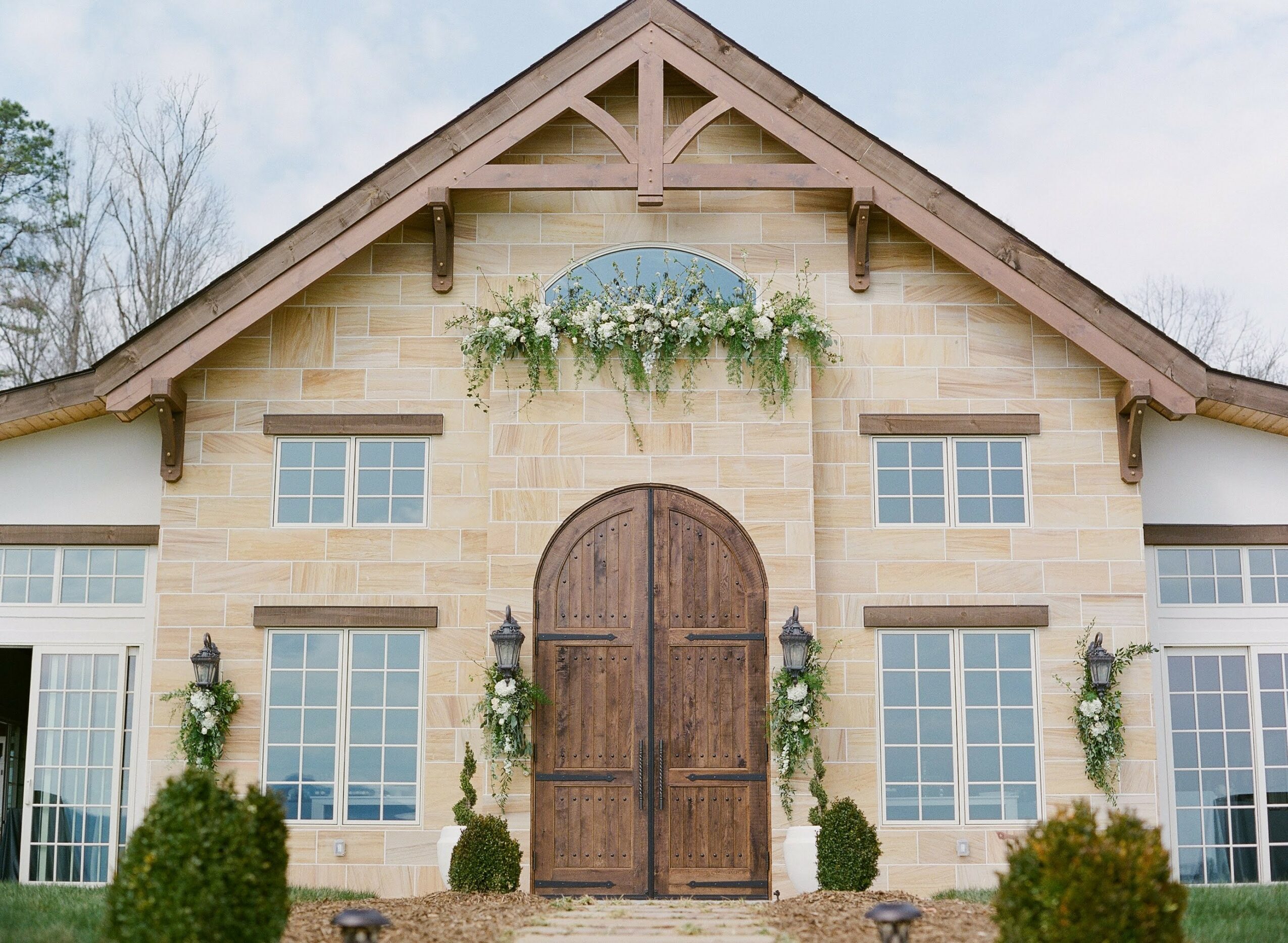 Front view of a house with a gabled roof, large windows, and a wooden arched door, decorated with greenery and plants.