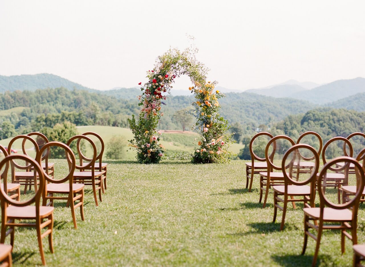Outdoor wedding setup with chairs facing a floral arch on a grassy field, mountains in the background.