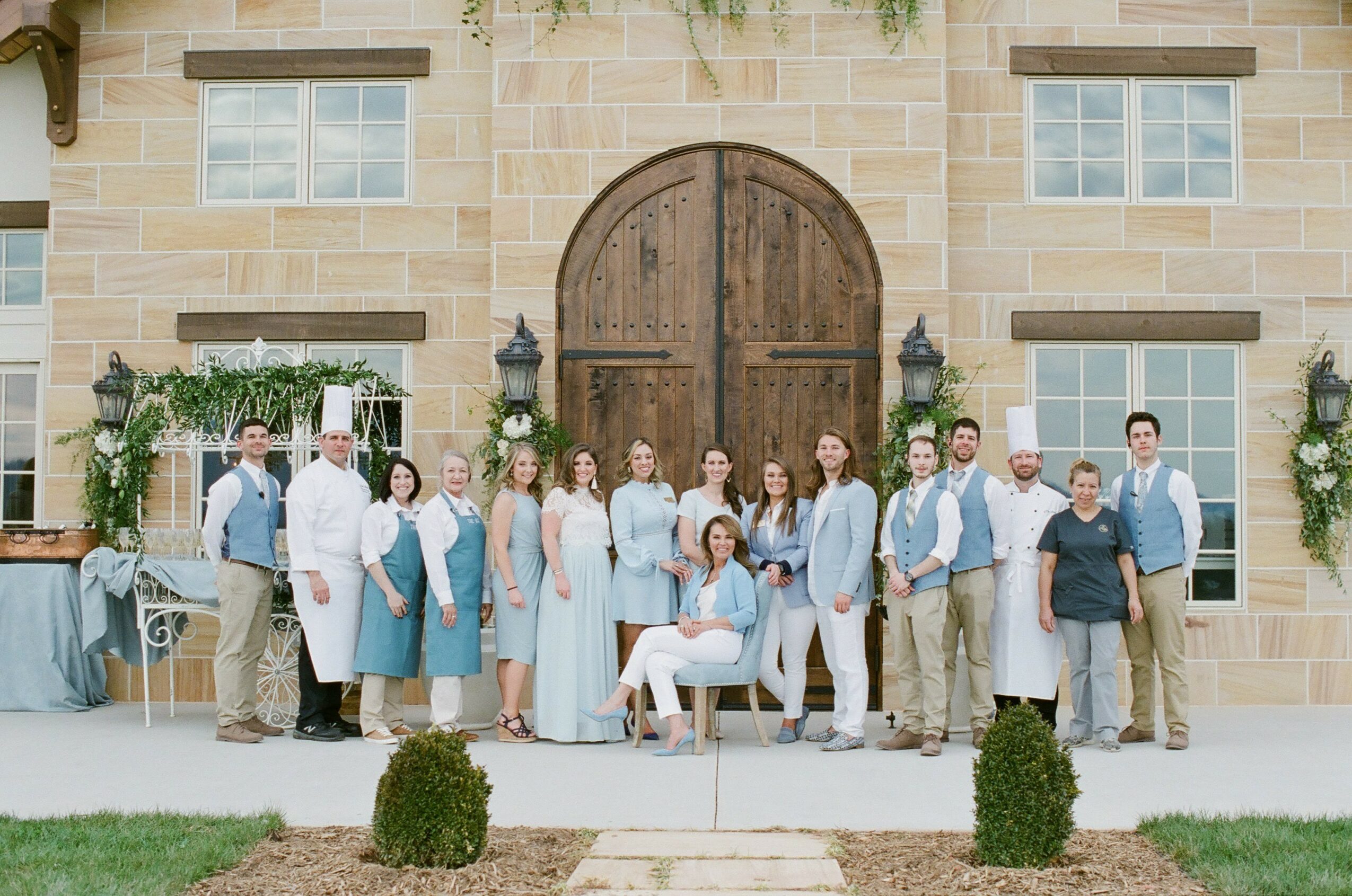 Group of people standing and sitting in front of a large wooden door on a stone building, with greenery and decorations.
