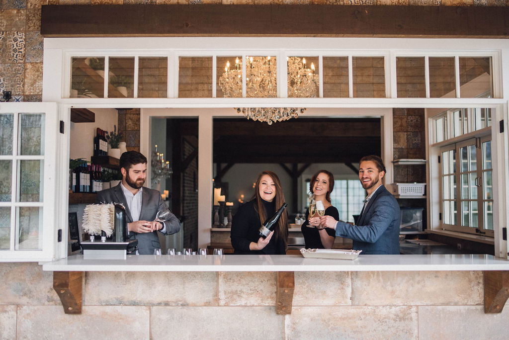 Three people standing behind a bar counter inside a room with large windows, smiling and holding drinks.
