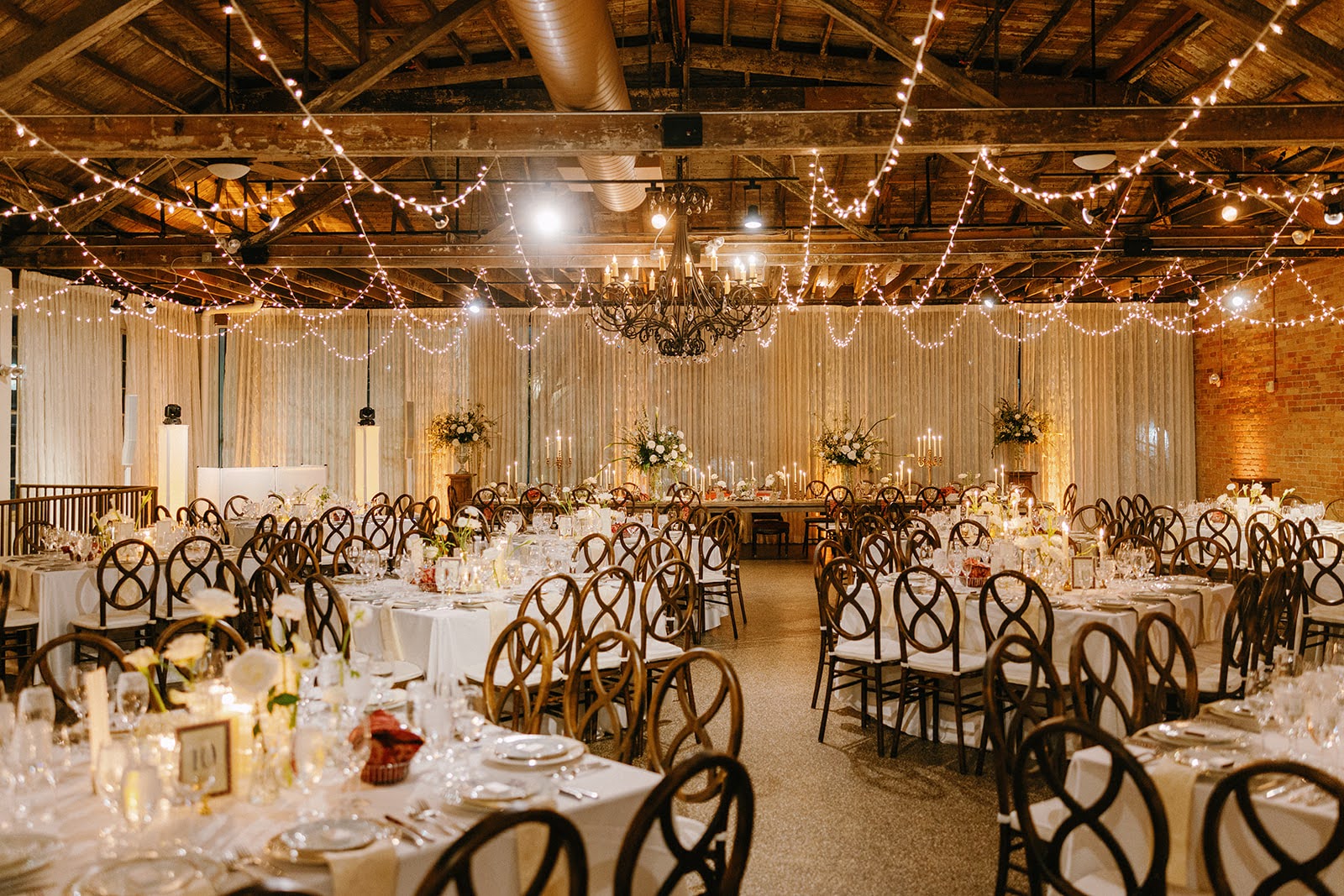 Elegant event space with round tables, chairs, string lights, and a chandelier, decorated for a celebration or wedding reception.